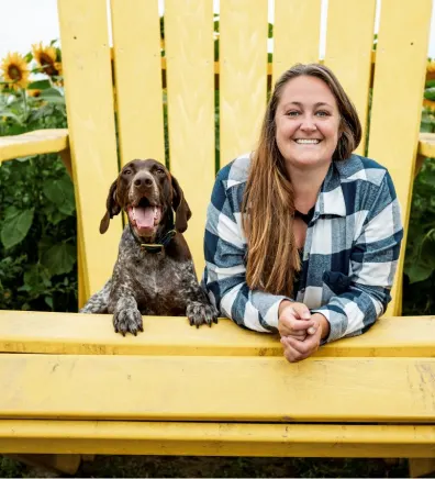 Nicole in a plaid shirt laying on a giant yellow lawn chair with a smiling dog.